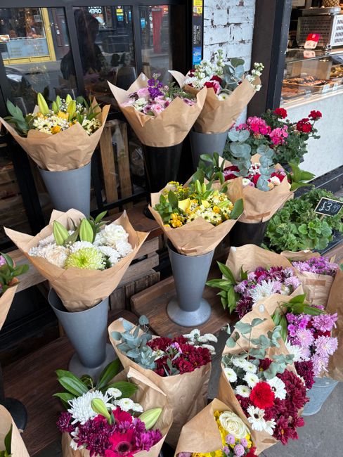 A display of various freshly cut flower bouquets arranged in tall, elegant grey and black vases outside a flower shop, with some bouquets wrapped in brown kraft paper. The arrangement includes a mix o