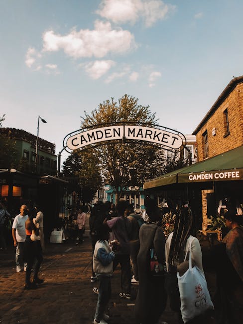 A vibrant scene at Camden Market with the iconic arched sign reading 'Camden Market' overhead amidst a lively crowd. In the foreground, there is a florist arranging a bouquet of fresh flowers with vis