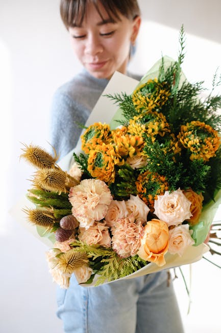 A young woman with light brown hair and a gentle smile holds a large, vibrant floral bouquet arranged with a mix of yellow, orange, and peach-colored flowers, including roses, carnations, and small da