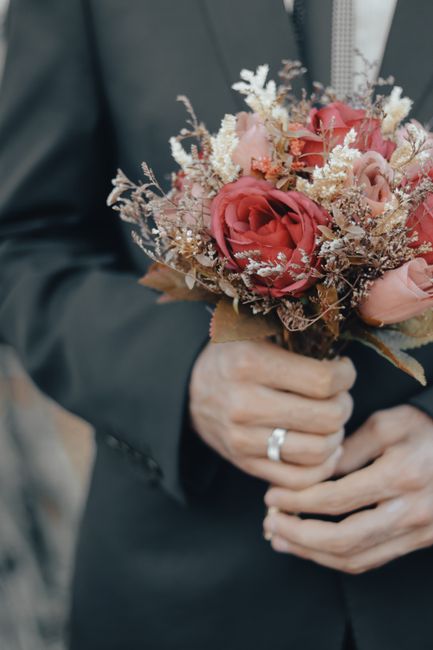 A person dressed in a dark suit holds a bouquet of fresh flowers, featuring large, velvety pink roses with delicate, light-colored dried foliage and smaller blooms, arranged in a compact, rounded styl