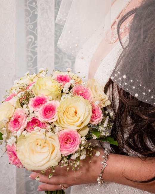 A bride holds a bouquet of fresh flowers featuring large cream roses and smaller pink roses, accented with delicate white baby's breath and green foliage. The bouquet is wrapped in soft white paper wi
